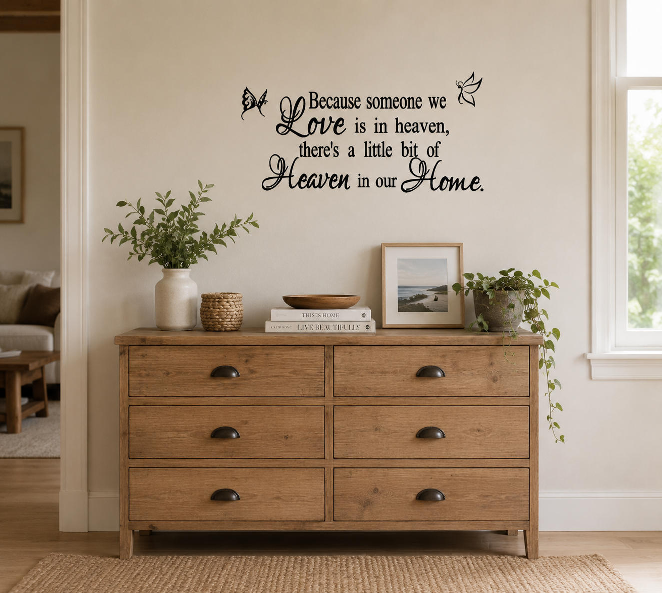Memorial wall decal featuring the quote “Because someone we love is in heaven” displayed above wooden drawers surrounded by family photo frames in a warm lounge room.